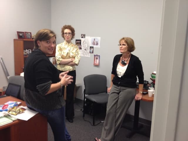 Three colleagues talking in a small branch office, one of them mid-sentence beside a desk, family photographs and a sketch taped to the wall behind them.