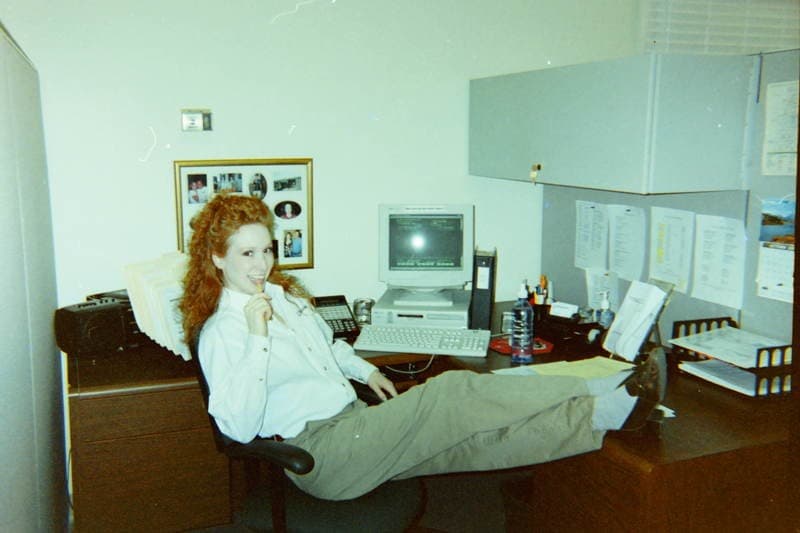 Nycole Rosen early in her staffing career, seated at her desk with her feet up beside a beige CRT monitor, paper files stacked behind her and printed schedules pinned to the cubicle wall.