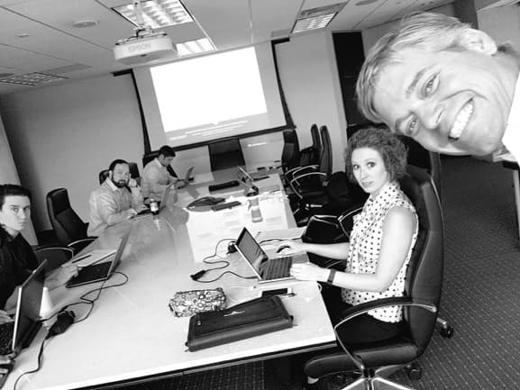 A black and white photograph of a project team working on laptops around a conference table, a lit projector screen at the front of the room, one colleague leaning into the frame from the right.